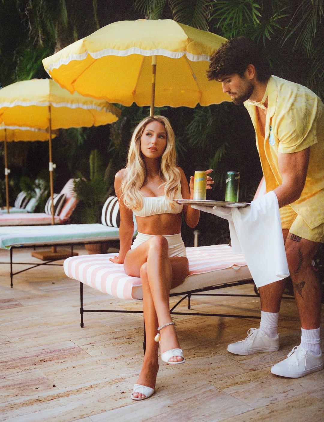 Alix Earle in a cream look beneath a yellow umbrella, with a server in a yellow uniform
